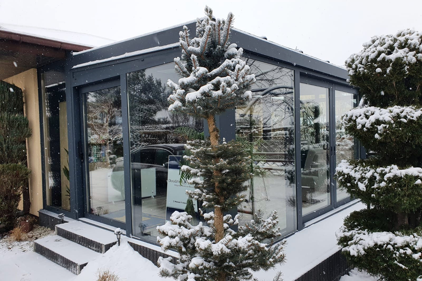 Antracietgrijze wintertuin met besneeuwde bomen ervoor en een dakvlak met sneeuwlast.