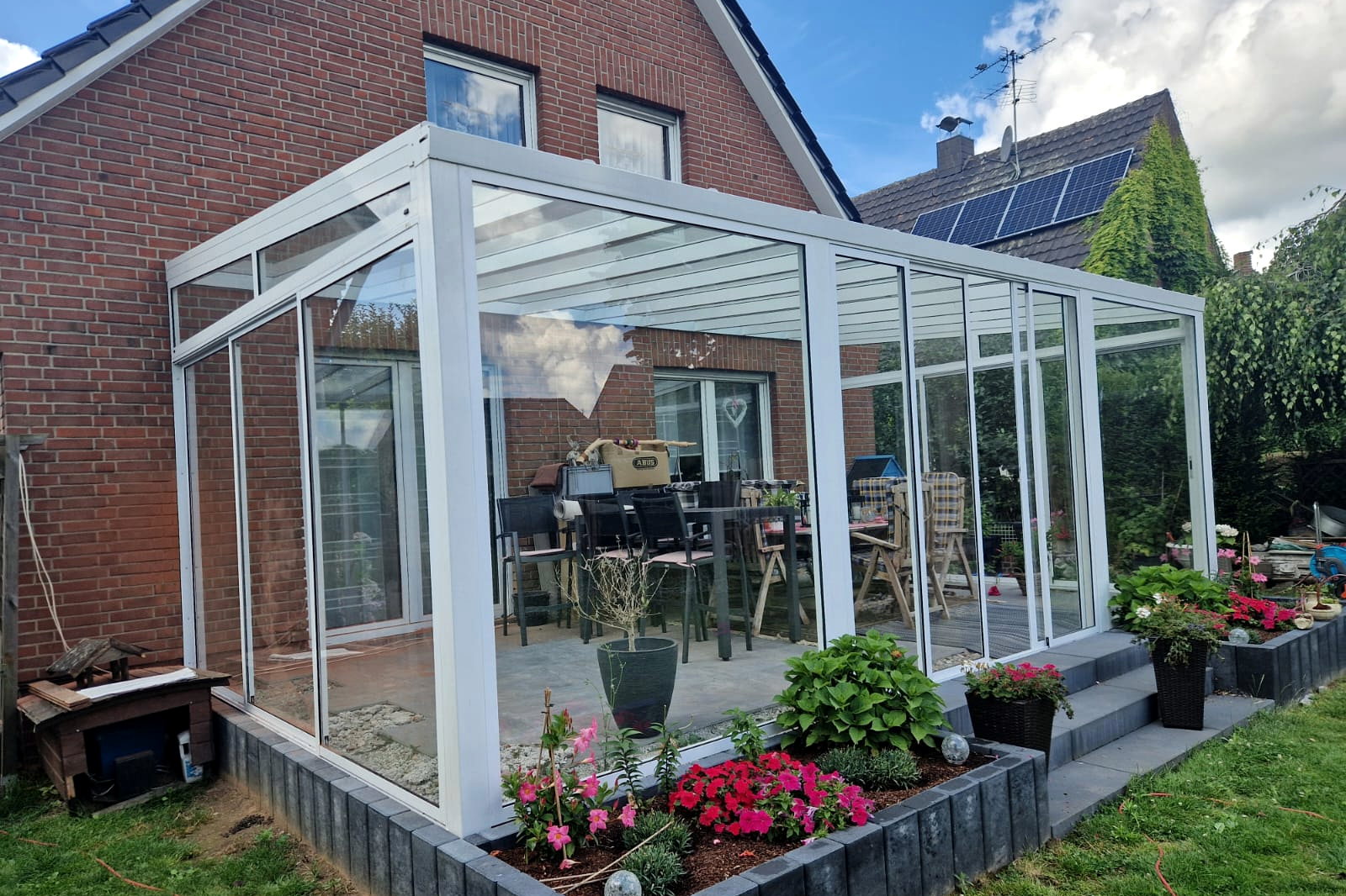 Witte wintertuin met glazen dak langs het terras van een bakstenen woning, met bloeiende border op de voorgrond.